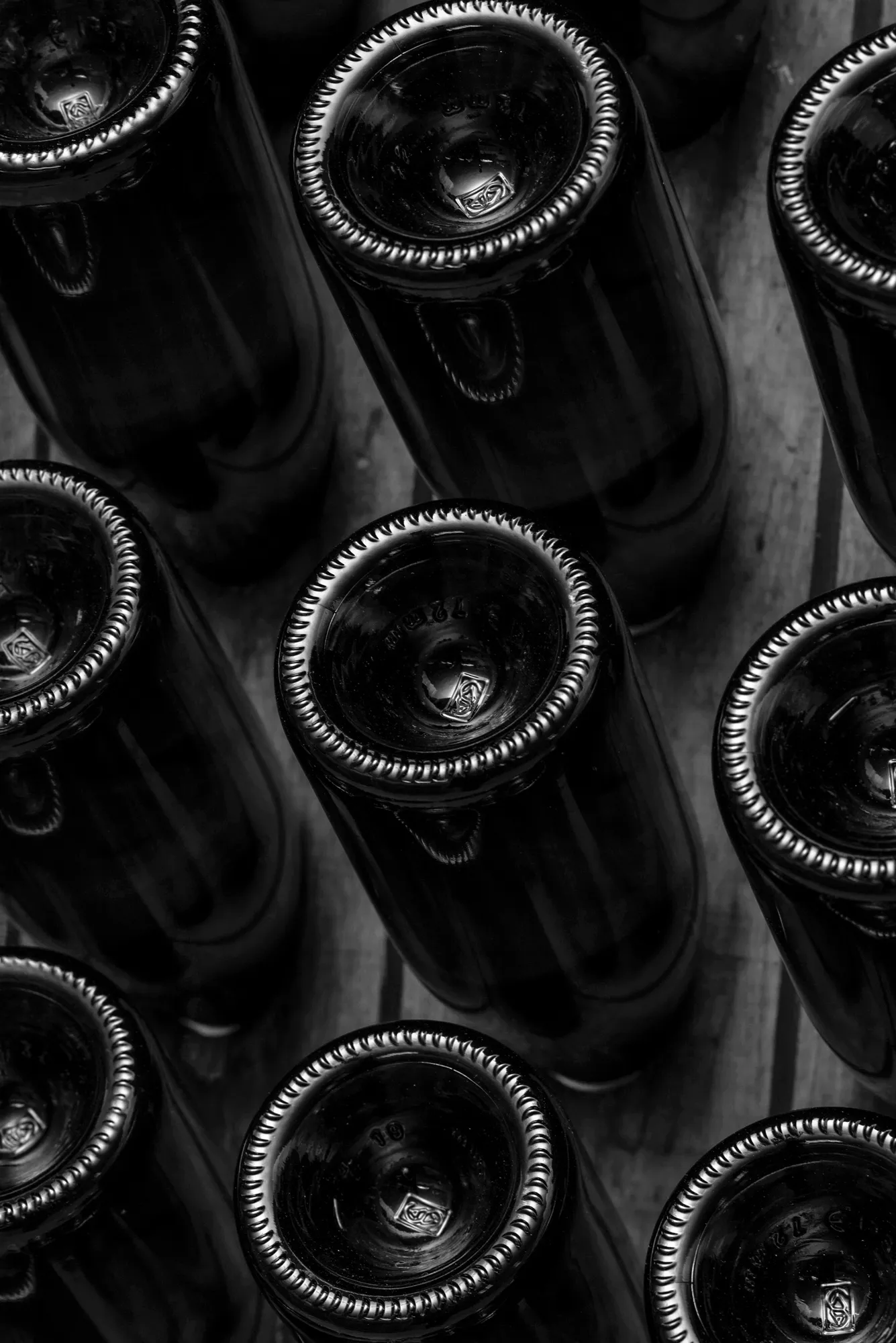 Dark wine bottles laid on their sides across wooden slats and shot from directly above, the knurled rims and embossed glass of their bases catching a hard raking light, in black and white